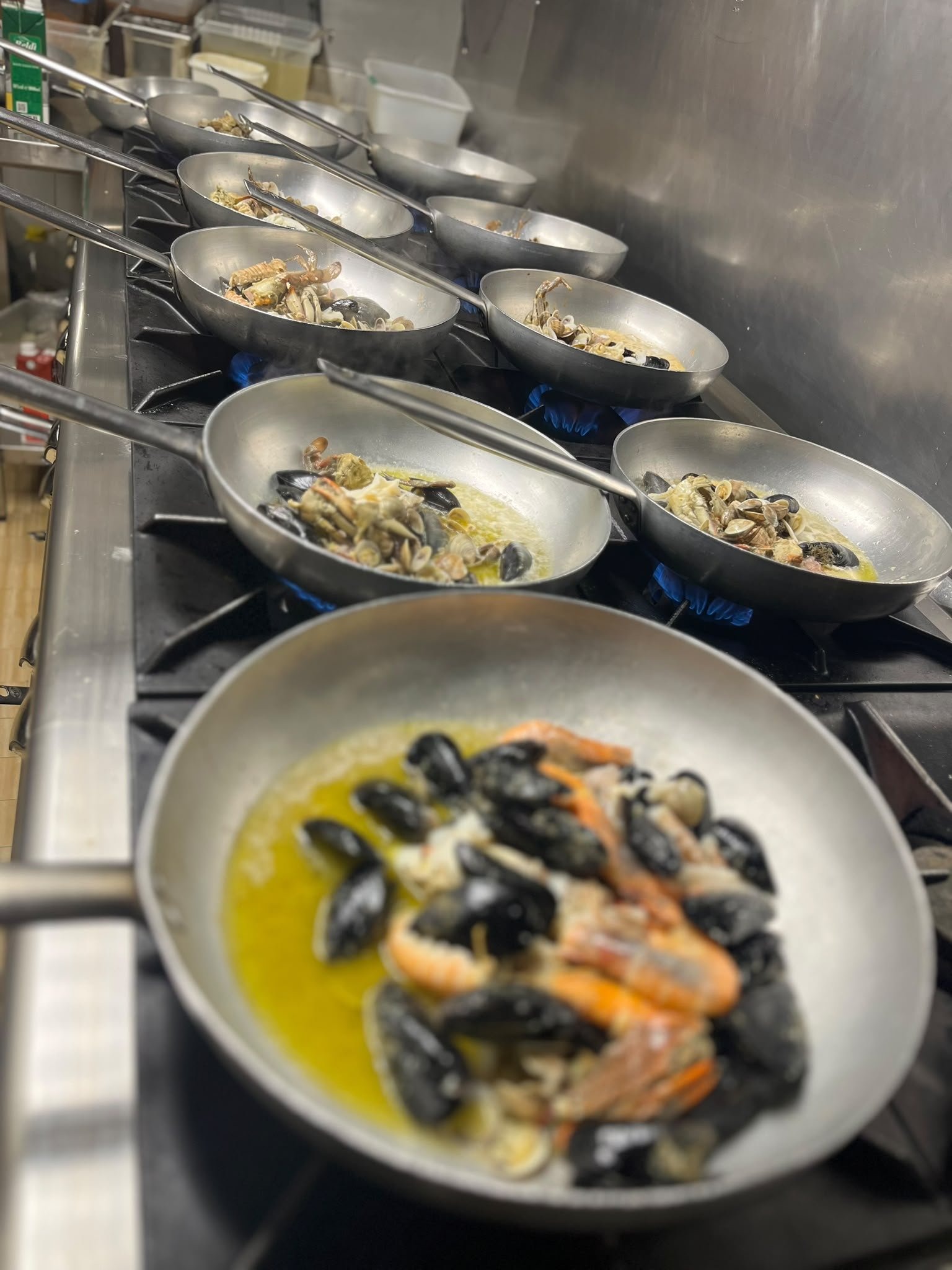 Padelle in alluminio sui fuochi a gas con cozze, scampi e vongole nell'olio, nella cucina del Ristorante della Rosa