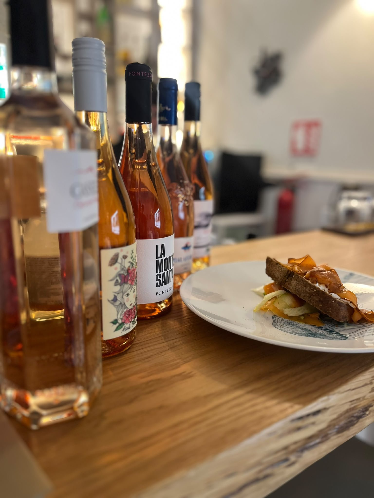 Bottiglie di vino rosato in fila su un bancone di legno accanto a un crostone con verdure servito su piatto bianco